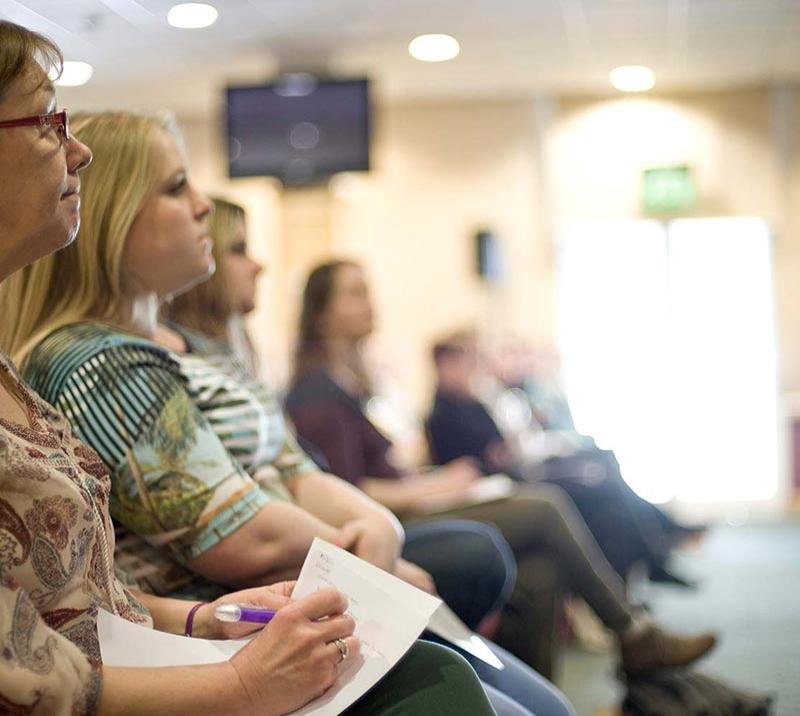 A seated crowd listen to an unseen speaker.  A lady in the foreground is taking notes.